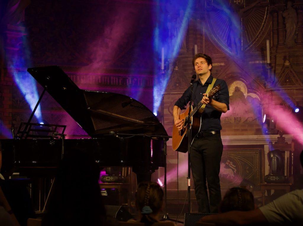 Portrait des Künstlers Lennert Schilgen mit Gitarre in bunt beleuchteter Kirche